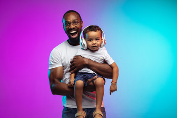 African-american father and son portrait on gradient studio background in neon. Beautiful male models in casual style, white shirt. Concept of human emotions, facial expression, sales, ad, family.