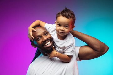 African-american father and son portrait on gradient studio background in neon. Beautiful male models in casual style, white shirt. Concept of human emotions, facial expression, sales, ad, family. - obrazy, fototapety, plakaty