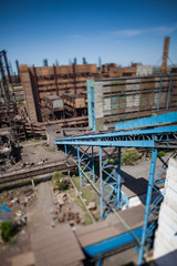 Fototapeta premium Old metallurgy factory buildings with blue belt conveyor and factory chimneys on blue sky. Birds-eye view and tilt-shift lens.