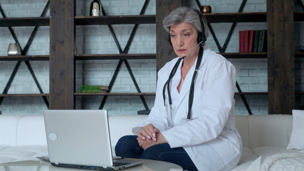 Naklejka na ściany i meble Middle aged woman wears a white coat and a headset while talking on a laptop computer using an online video call consultation app. Remote medical care for a remote patient
