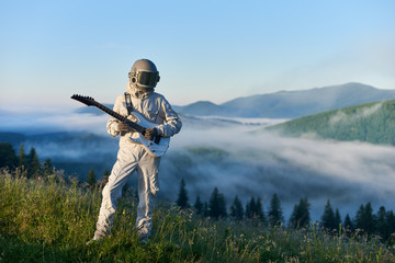 Cosmonaut in space suit and helmet playing guitar, standing on sunny green mountain glade in...