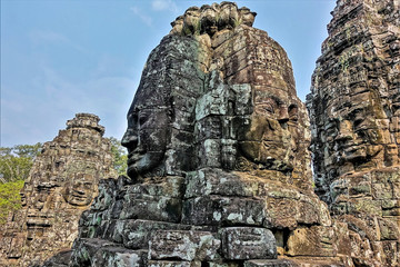 Fototapeta na wymiar Ancient mysterious temple of Bayon. Huge human faces carved from stone look at all directions. Strange smiles on their faces. The mystery of a bygone civilization. Background - blue sky. Cambodia. 