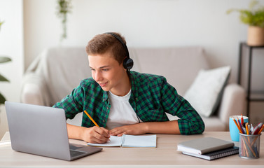 Joyful teen guy writing at notebook and looking at laptop