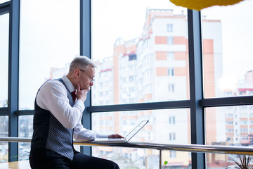 Fototapeta premium Adult male businessman, teacher, mentor working on a new project. Sits by a large window on the table. He looks at the laptop screen.
