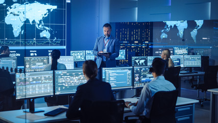 Senior Male Project Leader with Tablet Computer Makes an Announcement to the Team of Data Science Engineers. Telecommunications Control Monitoring Room with a Global Map on Big Screen.