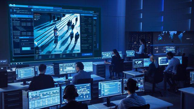 Team Of Professional Cyber Security Data Science Engineers Work On Surveillance Tracking Shot Of People Walking On City Streets. Big Dark Control And Monitoring Room With Computer Displays.