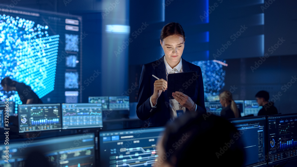 Poster Female Project Leader is Standing with Tablet Computer and Checking Work Data. Science Engineers Work Around Her. Telecommunications Control Monitoring Room with Neural Network on Big Screen.