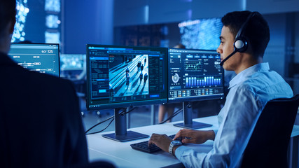 Confident Male Data Scientist Works on Personal Computer Wearing a Headset in Big Infrastructure Control and Monitoring Room. Surveillance Tracking Shot of People Walking on City Streets