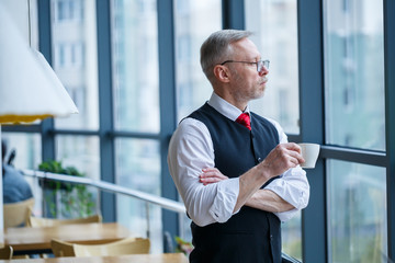 Fototapeta premium Smiling happy managing director thinks about his successful career development while standing with a cup of coffee in his hand in his office near the background of a window with copy space