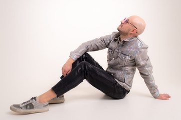 Stylish caucasian man without hair in grey shirt, black leather trousers and grey trainers with black sunglasses sits on the floor and poses for the camera isolated on white background