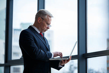 Fototapeta premium Smiling happy managing director thinks about his successful career development while standing with a laptop in his office near the background of a window with copy space
