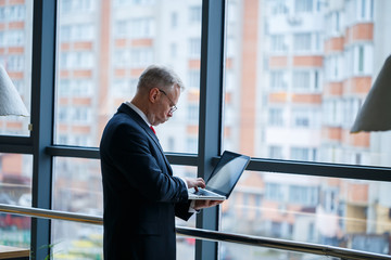 Fototapeta premium Smiling happy managing director thinks about his successful career development while standing with a laptop in his office near the background of a window with copy space