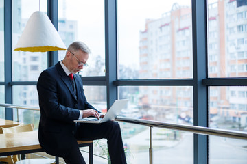 Fototapeta premium Smiling happy managing director thinks about his successful career development while standing with a laptop in his office near the background of a window with copy space