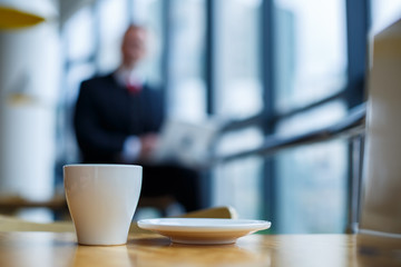 A white cup and saucer with hot aromatic coffee stands on a wooden table against the background of a sitting businessman