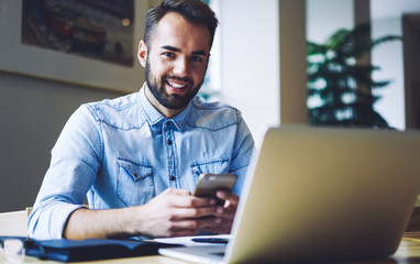 Obraz premium Half length portrait of smiling entrepreneur chatting on mobile phone with friends while looking at camera.Cheerful male student sitting at laptop computer while typing sms message on cellular