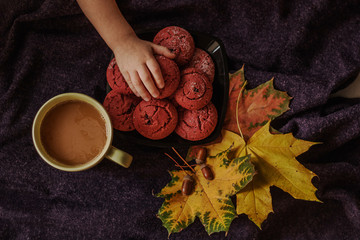 Children's hand reaching for a plate of cranberry cookies. Still life with a cup of coffee, autumn multi-colored maple leaves and acorns on the background of a purple plaid. Autumn concept. Top view