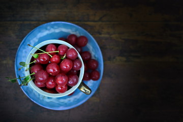 Blue cup on saucer with full red healthy ripe sweet cherry berries on old dark wooden background. Top view
