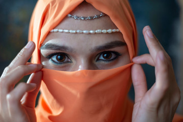 close-up portrait of a young Arab girl with beautiful eyes in an orange burqa, holding her hands...