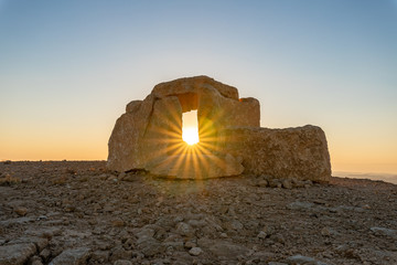 Rising  sun rays pass through a large stone dolmen at dawn in a public sculpture park in the desert, on a cliff above the Judean Desert near Mitzpe Ramon in Israel. © svarshik