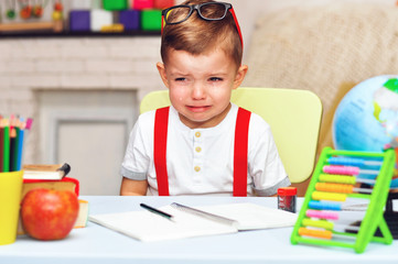 A little preschooler sits at a desk in a white T-shirt and red suspenders and crying.	