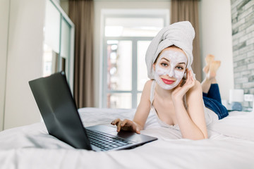 Young attractive woman after shower, with towel on head and mud facial mask, typing on laptop in bed, working or studying online at cozy bedroom at home. Working online, freelance concept