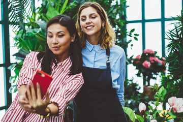 Positive multiracial female employee of floral store working in friendly atmosphere making selfie on telephone camera,asian woman taking picture with girl in apron satisfied with job in orangery