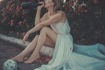 Fototapeta na wymiar Lonely bride thrown on the wedding day, drinking alcohol from a bottle, sitting on the pavement in a white dress. against the background of red rose flowers