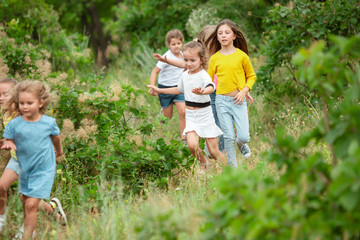 Fototapeta premium Best time. Kids, children running on green forest. Cheerful and happy boys and girs playing, laughting, running through green blooming meadow. Childhood and summertime, sincere emotions concept.