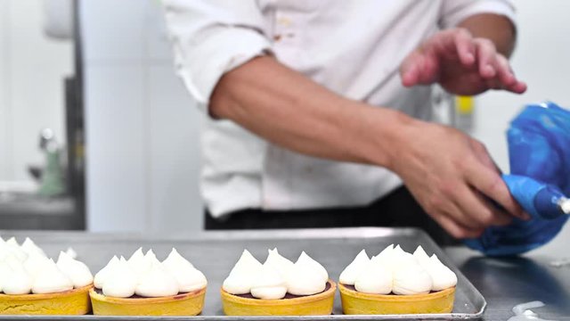 Pastry chef decorates biscuit tartlet with cream from pastry bag, close-up. Preparation of cake at commercial bakery with piping bag. High quality 4k footage.