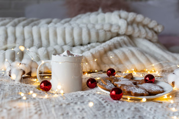 Christmas or New Year winter hot chocolate with marshmallows and gingerbreads, festive light garland and red Christmas tree toys on a white bed, selective focus.