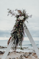 Top of white boho wedding arch wth flowers, roses, palm trees and greenery on the beach background. Outdoor ceremony decoration concept. Crete, Greece.