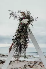 Spitze des weißen Boho-Hochzeitsbogens mit Blumen, Rosen, Palmen und viel Grün auf dem Strandhintergrund. Dekorationskonzept für Zeremonien im Freien. Kreta, Griechenland. © Jana Muranez