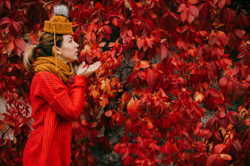 Portrait of a young beautiful caucasian female in a red sweater, black pants swabbed a scarf around her neck to keep warm in the cold autumn forest and blows to red leaves
