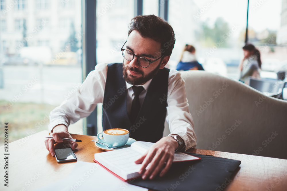 Poster young hipster guy in spectacles searching interesting news on smartphone device for writing in noteb