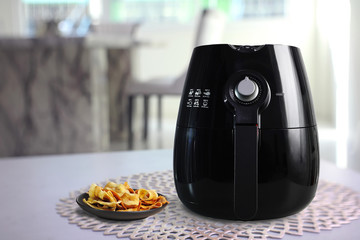 a black air fryer or oil free fryer appliance is on grey table in the dining room with deep fried banana chips in small black dish ( clipping path included )