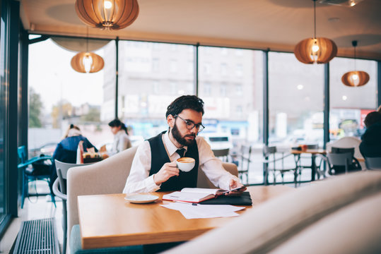 Caucasian hipster guy in trendy look reading sms message on modern smartphone from colleague while resting at cafeteria and enjoying cappuccino beverage, concept of technology and communication
