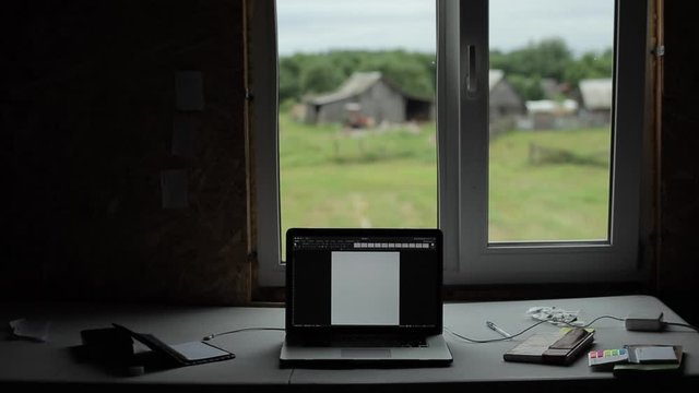 A laptop with books and notepads stands on the table by the window overlooking the rural landscape. The camera changes focus. Close-up