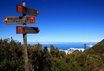 Three signs indicate hiking trails on the slopes of Pico Ruivo (Madeira, Portugal). In the background, the ocean and the blue sky merge.
