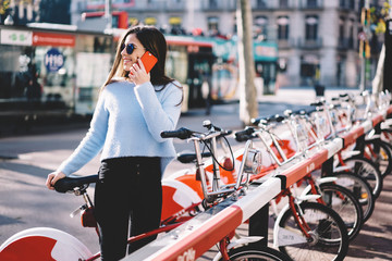 Fototapeta na wymiar Attractive young female calling to friend using modern smartphone arranging meeting and bicycle tour, trendy dressed hipster girl having phone conversation confirming rent enjoying active lifestyle