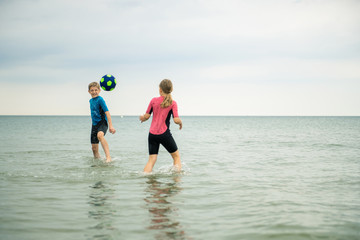 Two happy siblings children playing and jumping with ball in water in neoprene suits in sea