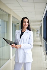 Fototapeta premium Young brunette woman, wearing white suit, holding black folder with documents,standing in white office building with huge windows.Businesswoman, getting ready for corporate meeting.Business relations