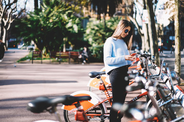 Attractive female hipster in sunglasses using smartphone for banking online renting bicycle for making touristic city tour, charming teen girl booking bike via mobile and 4g connection to internet