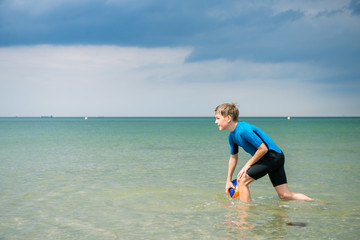 Happy handsome teen boy running and playing with ball in neoprene swimsuit in sea