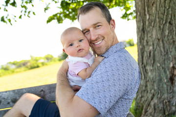 Father with baby girl standing outdoors by tree.