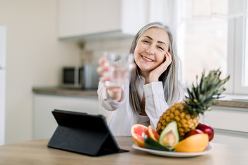 Joyful smiling Caucasian senior woman with long gray hair, showing glass of water to camera, while sitting at the table with fresh fruits and ipad in modern light kitchen. Focus on face