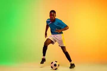 Leader. African-american male soccer, football player training in action isolated on gradient studio background in neon light. Concept of motion, action, ahievements, healthy lifestyle. Youth culture.