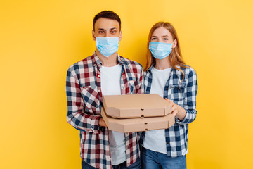 Fototapeta na wymiar Portrait of a couple, a man and a woman in checked shirts and medical protective masks on their faces, holding pizza boxes on an isolated yellow background