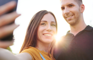 Happy millenial couple taking selfie outdoor in sunset - Smiling friends using smartphone in backlight - Sunny afternoon warm color tones with focus on girl holding phone