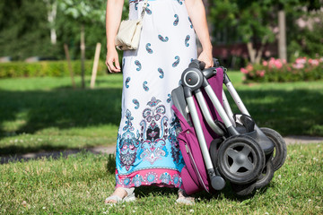 Unrecognizable woman holding folded baby pushchair, standing on green grass on lawn in sunny summer park