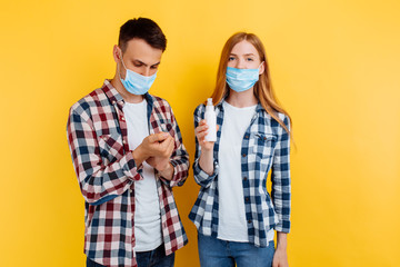 Young couple guy and girl in plaid shirts and medical protective face masks, disinfect their hands with antiseptic, standing on an isolated yellow background, coronavirus, health care